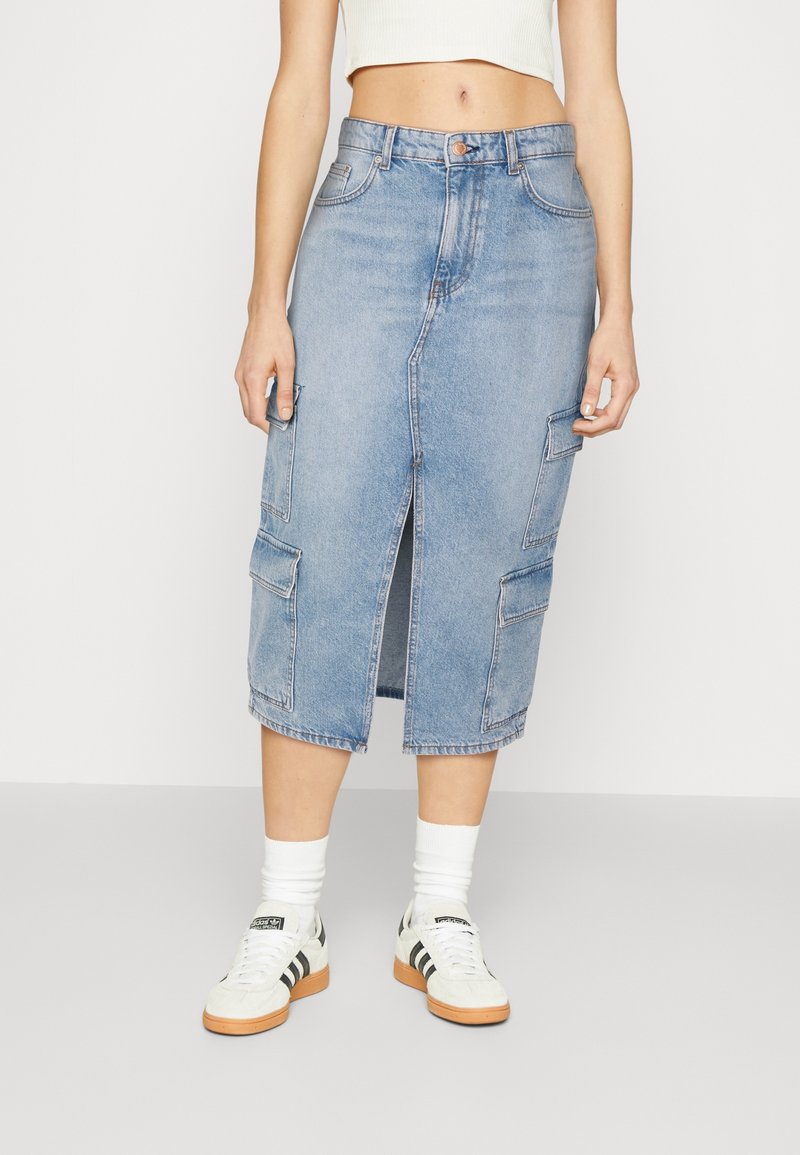 Femme portant une jupe midi en denim bleu clair avec une fente avant et des poches cargo, un crop top blanc, des chaussettes blanches et des baskets blanches.