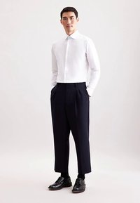 Witte blouse met lange mouwen en knopen, gecombineerd met marineblauwe geplooide broek en zwarte schoenen, staande pose tegen een lichte achtergrond.