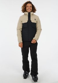 Beige und schwarze isolierte Jacke mit zwei Brusttaschen, hohem Kragen und Frontreißverschluss, kombiniert mit schwarzen Hosen und Skistiefeln.