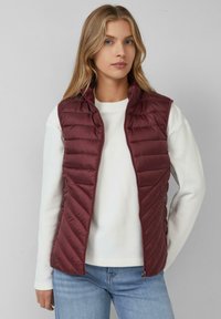 Weinrot gesteppte Weste mit Reißverschluss, Zickzacksteppung, hohem Kragen und Seitentaschen, über einem weißen Pullover getragen.