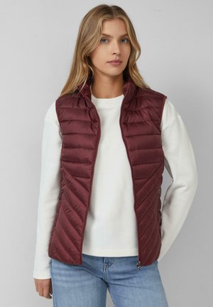 Gilet rembourré bordeaux avec fermeture éclair à l'avant, design en chevrons matelassé, col montant et poches latérales, superposé à un pull blanc.
