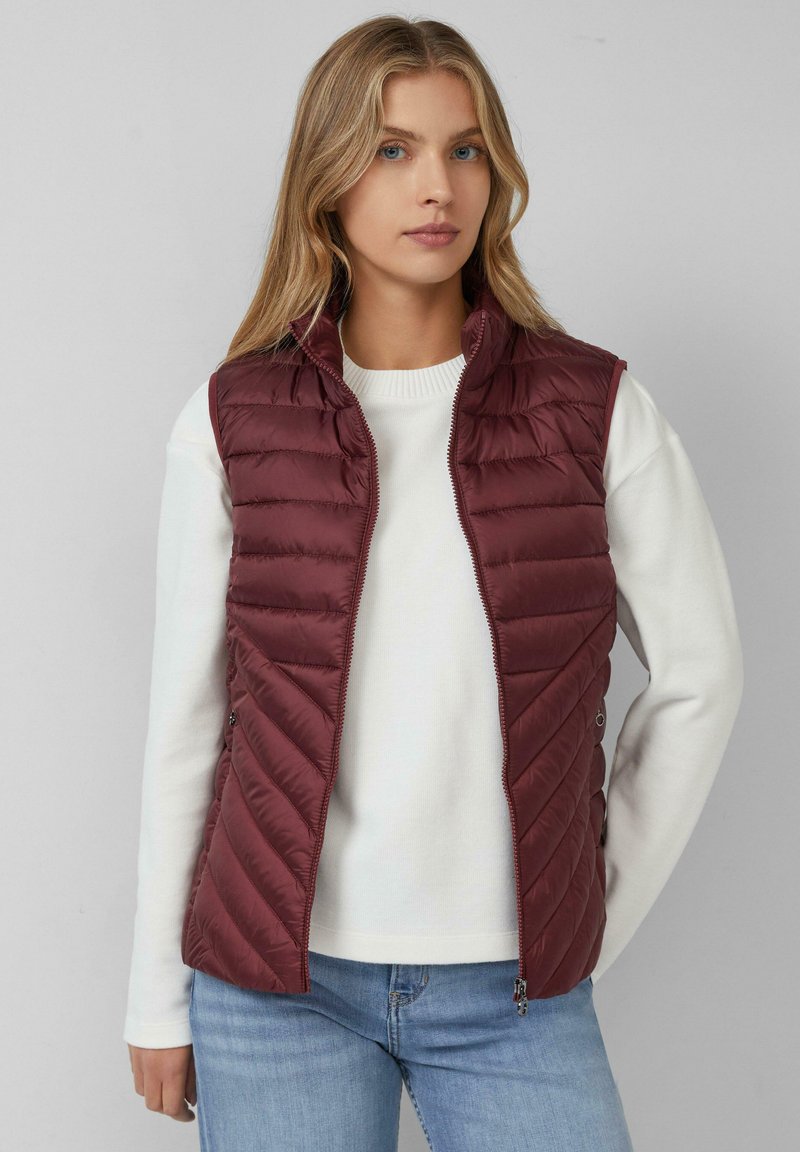 Weinrot gesteppte Weste mit Reißverschluss, Zickzacksteppung, hohem Kragen und Seitentaschen, über einem weißen Pullover getragen.