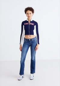 Mörkblå och vit kort zip-up jacka med "COSMIC"-logotyp, tillsammans med ljusblå utsvängda jeans och vita sneakers. Minimalistisk design.