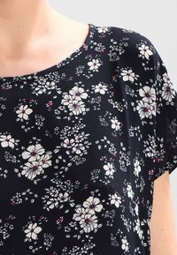 Blouse à fleurs bleu marine à manches courtes ; présente des fleurs blanches et bordeaux sur un tissu texturé ; encolure arrondie et coupe décontractée.