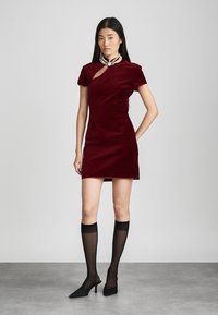 Robe en velours bourgogne à manches courtes, avec une découpe à l'épaule. Associée à des bas noirs en cuir montant jusqu'à la cuisse et des escarpins noirs à bout pointu.