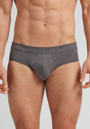 Mandlig torso iført tætsiddende mørkegrå briefs, der viser veldefinerede mavemuskler og let behårede øvre ben mod en ensfarvet baggrund.