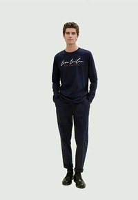 Marineblaues Langarmshirt mit weißem Schriftzug-Logo, kombiniert mit passenden dunklen Hosen und schwarzen Schuhen. Legere Passform und glatte Stoffstruktur.