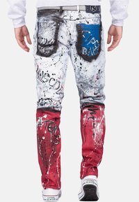 Jeans in denim con una base bianca e macchie di vernice nera e rossa, dettagli strappati, una tasca blu e accenti testuali in stile graffiti.