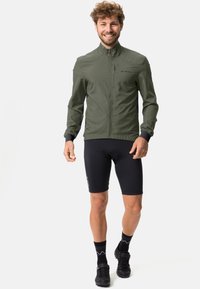 Olivgrön lättviktsjacka med ståkrage, dragkedja fram och sidopaneller, bärs med svarta cykelshorts och skor.