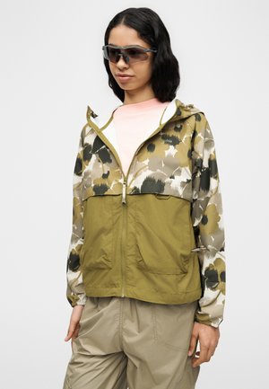 Ženska, ki nosi temna sončna očala, zeleno-bež jakno z zadrgo in cvetličnim vzorcem ter bež hlače, stoji pred enobarvnim svetlim ozadjem.