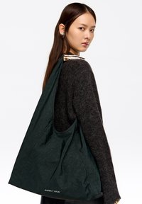 Duża zielona torba typu tote wykonana z teksturowanej tkaniny o miękkim, opadającym kroju. Posiada nadrukowaną metkę na środku u dołu torby.