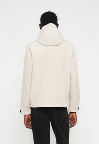 Homme portant une veste à capuche beige et un jean noir, de dos, sur un fond blanc uni.