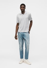 Άνδρας που φορά ανοιχτό γκρι πόλο με κοντά μανίκια, ανοιχτό μπλε τζιν και λευκά αθλητικά παπούτσια, στέκεται μπροστά σε ένα απλό λευκό φόντο.