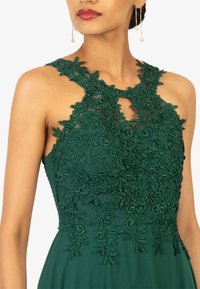 Groene jurk met bloemenkant overlay, doorzichtige halslijn en ingewikkelde borduursels. Kenmerkt zich door een mouwloos ontwerp en een uitsnijding bij de borst.