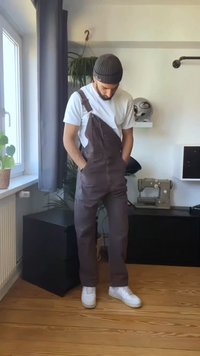 Homem vestido com macacão escuro, camiseta branca e gorro está dentro de casa perto da janela com as mãos nos bolsos, com uma máquina de costura e uma planta por perto.