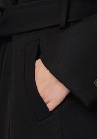 Czarny wełniany płaszcz o miękkiej fakturze, z kieszenią na lewym rękawie. Zapinany na pasek z guzikiem na rękawie.