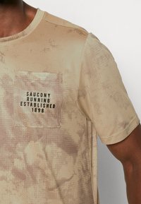 Bežové tričko s krátkým rukávem se strukturovaným vzorem a kapsou na levé straně hrudi, na které je potištěno „Saucony Running Established 1898“ černou barvou.