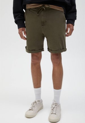 Homme portant un short vert olive à ourlet roulé, des chaussettes blanches mi-mollet, des baskets écrues et un haut noir à manches longues, debout sur un fond neutre.