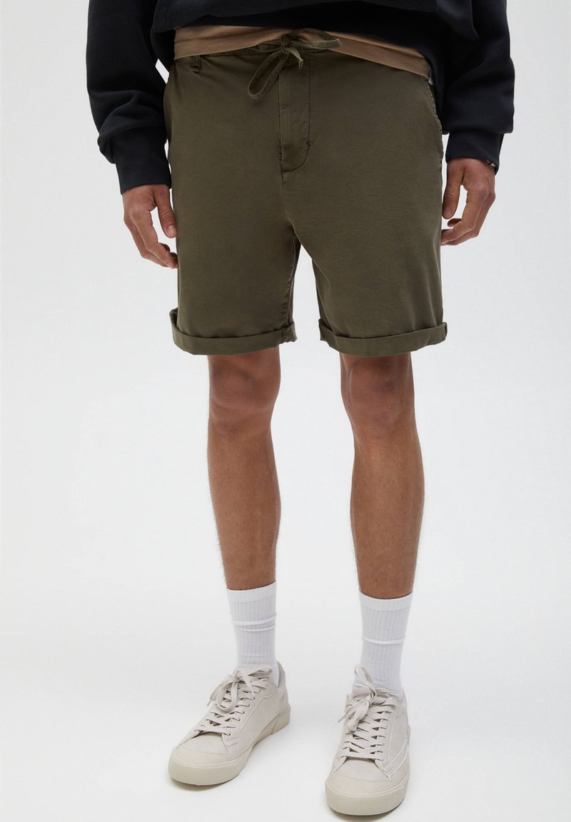 Homme portant un short vert olive à ourlet roulé, des chaussettes blanches mi-mollet, des baskets écrues et un haut noir à manches longues, debout sur un fond neutre.