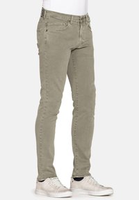 Jeans slim-fit verde oliva chiaro realizzati in cotone morbido, con tasche frontali e chiusura con zip, abbinati a sneakers bianche.