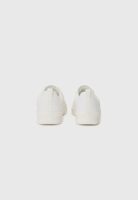 White canvas sneakers with rubber soles, featuring a sleek design, elastic sides, and looped heel tabs for easy wear.