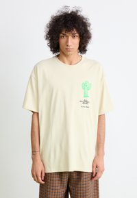 Kremfarget oversized t-skjorte med korte ermer, med et grønt kaktusmotiv og tekst på venstre brystområde. Avslappet design.