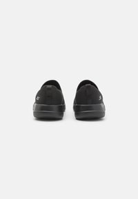 Zapatillas negras tipo slip-on con un upper de malla texturizada, suela de goma lisa y una lengüeta en el talón para facilitar el uso. Branding mínimo en el lateral.