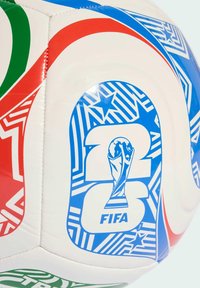 Tuvumā redzama balta futbola bumba ar krāsainiem sarkaniem, zaļiem un zilajiem ģeometriskiem rakstiem un FIFA Pasaules kausa trofejas emblemu.
