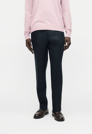 Personne portant un pantalon noir ajusté, des mocassins marron foncé et un pull à manches longues rose clair sur un fond blanc uni.