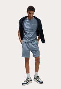 Helles blaues Baumwoll-T-Shirt mit einer Brusttasche, kombiniert mit hellblauen Shorts. Darüber trägt die Person eine schwarze Jacke über den Schultern und sportliche Sneaker.