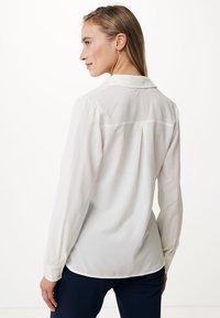 Camicia bianca a maniche lunghe con colletto, caratterizzata da una piega sul retro e polsini con bottoni, realizzata in tessuto leggero. Texture liscia e vestibilità sartoriale.