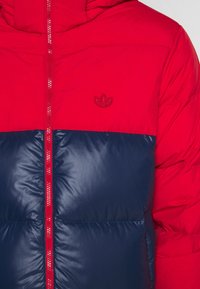 Röd och marinblå puffjacka med dragkedja, horisontell färgblockdesign och broderad logotyp på bröstet. Sl�ät textur.