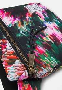 Jasny, wielokolorowy worek Herschel z zamkiem błyskawicznym i skórzaną pętelką w kolorze czarnym na białym tle, ukazujący teksturę materiału i detaliczne szwy.