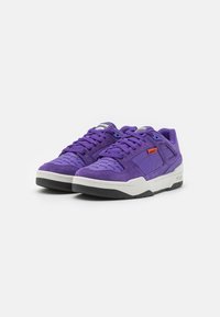 Lilla suede sneakers med en struktureret overdel, gummisål og kontrastfarvet hvid mellemsål. Har et rødt mærke og logo detaljer.