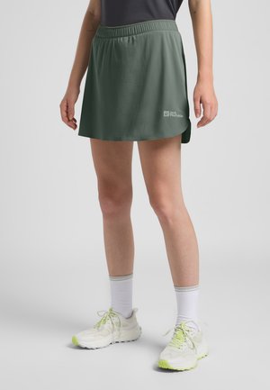 Grüner Sportrock mit einem elastischen Bund, ausgestattet mit einem seitlichen Schlitz und einem kleinen Logo. Kombiniert mit weißen Laufschuhen und grauen Socken.