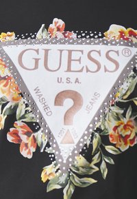 Sort stof med et trekantet logo med blomsterdesign, rhinsten detaljer og tekst: "GUESS U.S.A. WASHED JEANS" med et spørgsmålstegn.