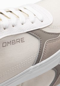 Beżowo-kremowy sneaker z teksturowanym cholewką z płótna, gładkimi skórzanymi akcentami, płaskimi białymi sznurowadłami oraz logo "OMBRE" w jasnoszarym kolorze.