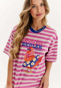 Růžové a bílé pruhované tričko s modrým výstřihem, se barevným grafikou kuřete v vzorovaných šatech. Text zní "LOKÁLNÍ OBCHOD S VEJCI".