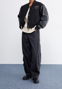 Zwarte varsityjack met leren mouwen, voorzien van geborduurde logo's. Gecombineerd met losse donkere jeans met cargopockets en zwarte laarzen.