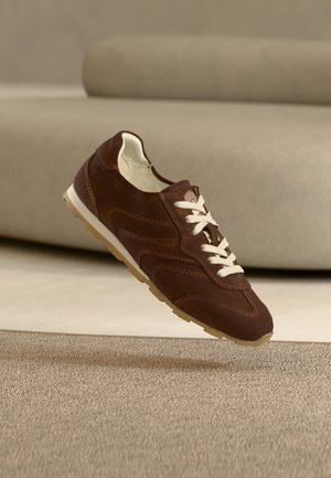 Baskets en cuir marron avec des lacets blancs suspendues au-dessus d'un tapis beige, canapé beige flou en arrière-plan.