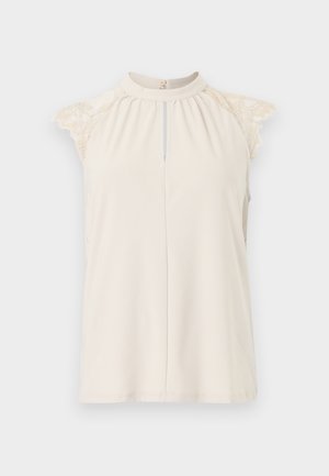 Αμάνικη κρεμ μπλούζα με ψηλό γιακά, άνοιγμα μπροστά και δαντελωτές λεπτομέρειες στους ώμους, πάνω σε απλό ανοιχτό φόντο.
