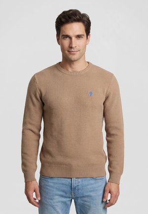 Uomo che indossa un maglione beige strutturato con un piccolo logo blu a forma di albero sul petto e jeans azzurri, in piedi su uno sfondo semplice.