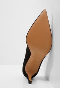 Chaussure à talon haut noire pour femme à bout pointu affichée semelle vers le haut, montrant une semelle en cuir beige et un talon fin sur fond blanc.