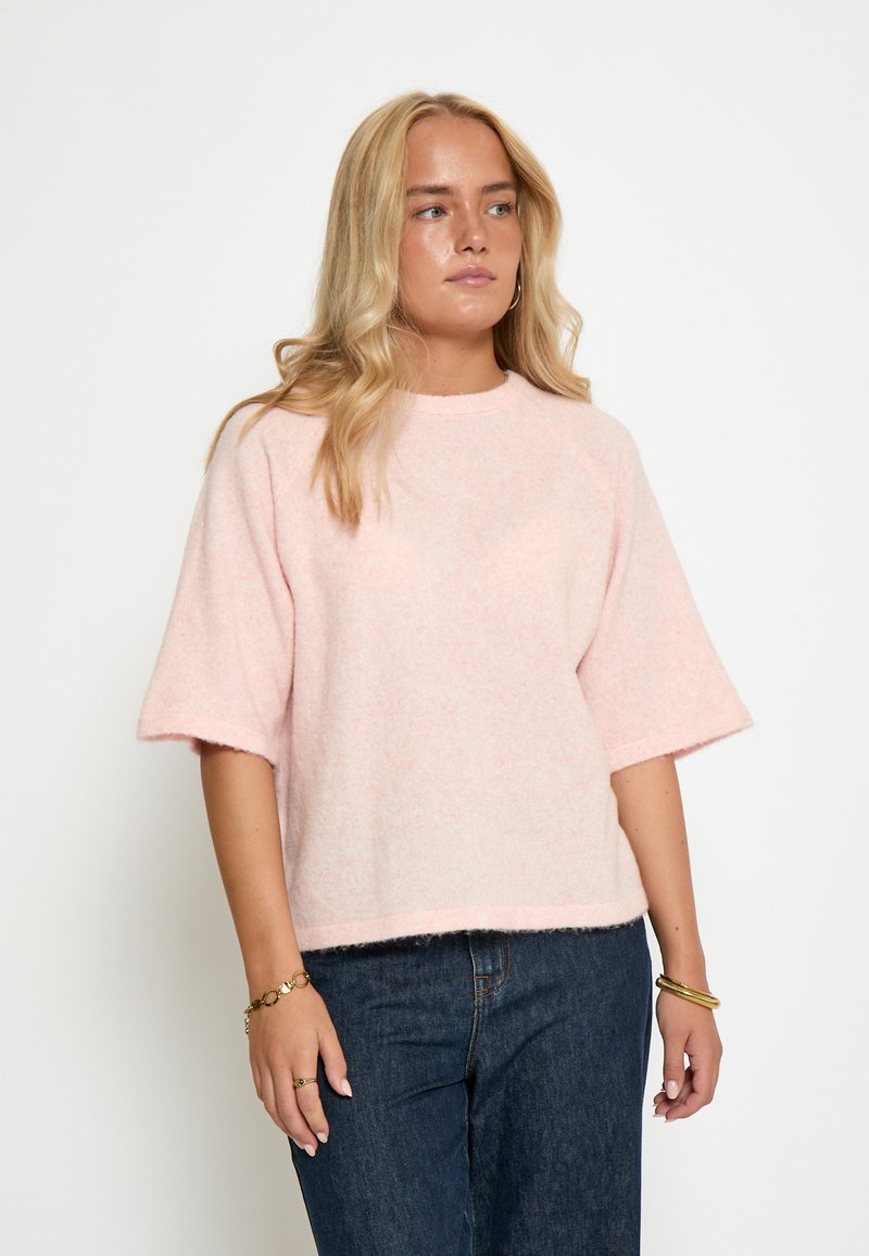 Gaiši rozā, pūkaina džemperis ar īsām piedurknēm, apaļu kakla izgriezumu un mīkstu tekstūru. Kombinēts ar tumšām džinsu biksēm.