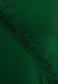 Dunkelgrüner Stoff mit gerippter Textur, der entlang einer Seite mit einem diagonalen Rüschensaum verziert ist, was einen visuellen Kontrast und Detail schafft.