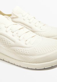 Lätta, helvita sportskor med andningsbar meshtopp, platta snören och strukturerad gummisula. Utformade för komfort och andningsförmåga.