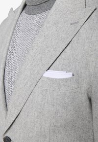 Grå blazer lavet af tekstureret stof, med en klassisk krave, sidelommer og et hvidt lommetørklæde i brystlommen.