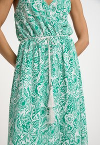 Grön och vit blommönstrad klänning med v-ringning, elastisk midja med en vit flätad snörning och en dekorativ tofs.