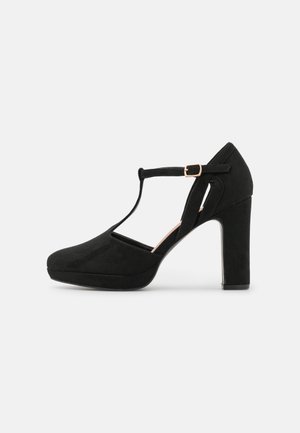 Zapato de tacón alto en ante negro con correa en T, puntera redondeada, suela con plataforma y correa ajustable con hebilla dorada sobre un fondo liso.