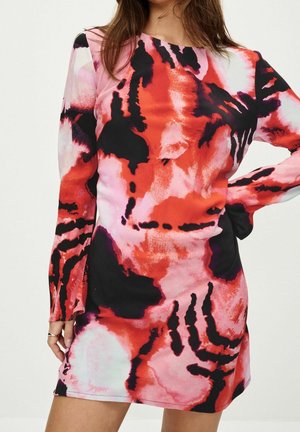 Femme portant une robe courte à manches longues avec des motifs abstraits rouges, roses, noirs et blancs, debout avec une main sur la hanche.